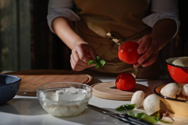 Close-up of housewifes hands. Chef holding ripe tomato and basil leaves while preparing meal in a rustic kitchen. Raw ingredients on chopping board and fresh feta cheese on glass bow on white table