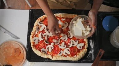 Top view of a pizzaiolo, the chef puts fresh mozzarella cheese balls on top of the rolled out dough smeared with tomato sauce and organic champignons while making delicious pizza. Italian traditions