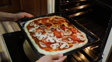 Details: Hands of a housewife, chef putting a baking sheet with raw vegetarian pizza into the oven. Preparing delicious healthy food at home. Italian traditional cuisine