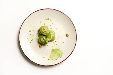 Flat lay. Food still life of three frozen green balls of pistachio and lime ice cream sorbet,sprinkled with pistachio nuts, melting on a plate, isolated on white background with copy space for text.