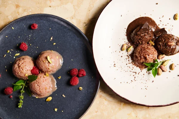 Flat lay. Frozen balls of refreshing raw vegan ice cream topped with raspberries and lemon basil leaves and chocolate dairy free sorbet, melt on plates, against a marble table. Still life with food