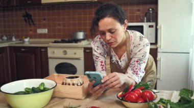 Beautiful woman, housewife in chefs apron, using smartphone, looks through a recipe for pickled vegetables on websites, standing at home kitchen background. Pickling vegetables. Canning concept