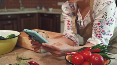 Soft focus on a smartphone in the hands of a woman, housewife wearing a chefs apron, looking through recipe of marinating and pickling seasonal vegetables at home kitchen. Close-up. Family traditions