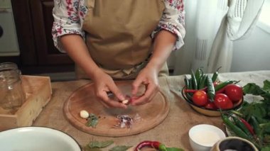 Close-up. Hands of a housewife in a chefs apron peeling fresh garlic, chopping it on a wooden board, standing at a table with organic vegetables and ingredients for making marinade in a home kitchen