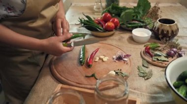 Close-up. Hands of a housewife in a chefs apron, cutting chili peppers on a chopping wooden board, standing at a table with organic vegetables and ingredients for making marinade in a home kitchen
