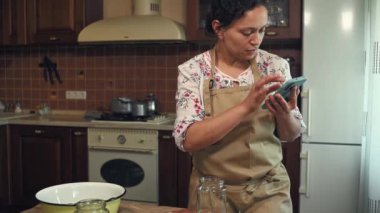 Multiethnic woman, housewife in a beige chefs apron, looking through the smartphone and preparing canned cucumbers according to a recipe from the Internet, in a cozy home kitchen. Pickling. Canning.