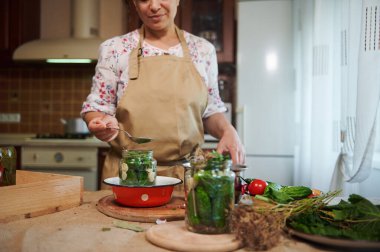 Focus on a jar of fresh cucumbers on a blurred background of a woman, housewife in a beige kitchen apron, pouring marinade into a jar while making pickled cucumbers. Marinating, canning, pickling.