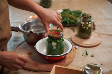 Close-up of a housewifes hand, putting a sterile lid on a glass jar of freshly pickled organic cucumbers. Homemade canned food. Preparing marinated delicacy for the winter season. Pickling.