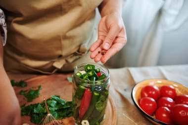 Details: Womans hand adding peppercorns in glass jar for canning, filled with stacked fresh chili peppers while making homemade spicy preserves at home kitchen