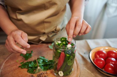 Cropped view of housewifes hand adding fragrant culinary herbs and peeled fresh garlic cloves in freshly marinated red and green chili peppers. Close-up pickling, canning, marinating. Family recipe