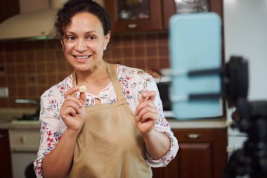 Pretty woman, housewife, wearing a beige chefs apron holding a peeled garlic clove and an umbrella dill, smiles looking at camera, mounted on tripod while recording video blog about seasonal pickling