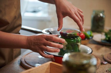 Details: housewifes hands cover with a sterilized lid a glass jar with freshly pickled organic cucumbers. Canning, marinating, pickling concept. Preparing homemade marinated delicacies for the winter