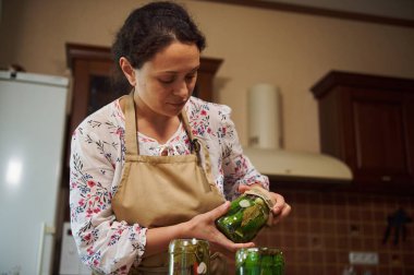 Portrait of a pleasant multiethnic woman, housewife in chefs apron cooking homemade delicacies for the winter, pickled cucumbers and different vegetables in the cozy wooden rustic home kitchen