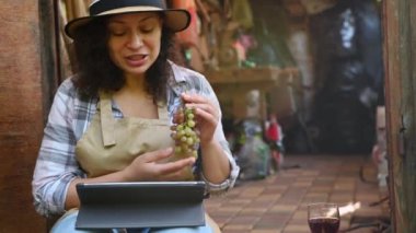 Close-up of a beautiful woman in a beige apron and straw hat, sitting on the doorstep of her workshop, presenting an assortment of grapes grown in her own vineyards while having a video call on tablet