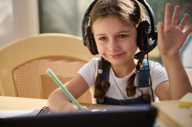 Close-up portrait of an European child girl, schoolgirl, smart elementary school student, wearing wireless audio headset, smiling sweetly while watching online video lesson, studying in online school
