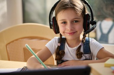 Headshot. Adorable Caucasian cute girl, schoolgirl, first grader wearing wireless headphones, sweetly smiling while looking at camera, holding pencil and browsing online video lesson on digital tablet