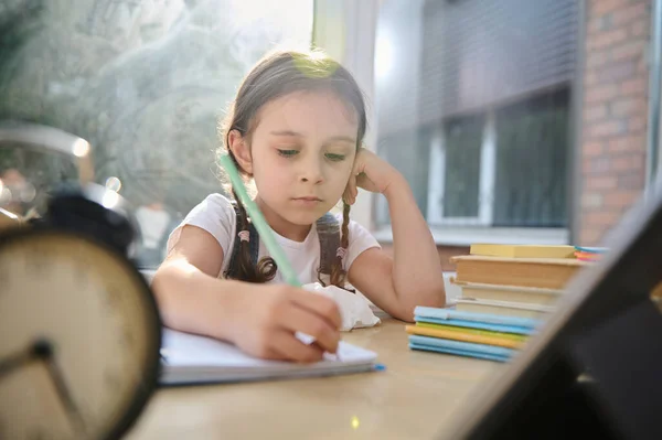 Sunbeams fall on the classroom through large panoramic windows while a charming schoolgirl, primary student, first grader does her homework, studies the writing, remotely from home in online school