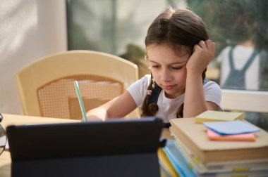 Charming serious schoolgirl studying remotely from home, learning the writing lesson, watching online language courses on a digital gadget. The benefit of distance education. Knowledge. School concept