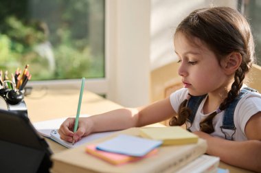 Smart elementary age school girl doing her homework with digital tablet at home, learning the writing by watching a broadcasted online lesson. Education and distance learning for kids. Homeschooling