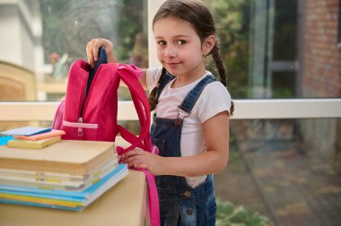 Caucasian student of primary school, with two pigtails, wearing a white t-shirt and blue denim overalls, smiles at camera while folding her pink backpack, happy to be a first grader. Back to school