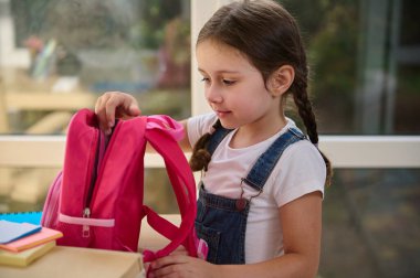 Close-up of European smart schoolgirl with two pigtails, in white T-shirt and blue denim overalls, stands at a table on the veranda of the house and puts her school supplies into a pink school bag