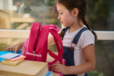 Inspired European first grader, schoolgirl in denim overalls, folding notebooks and school supplies in a pink knapsack, getting ready for the first grade of elementary school, early in the morning