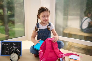 Adorable Caucasian student of primary school, schoolgirl, first grader wearing white t-shirt and blue denim overalls, sitting on a table near an alarm clock and chalk board, folding her pink backpack