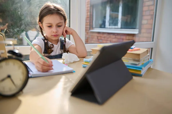 Virtual distance learning. Little Caucasian child, first grader, school girl, pupil kid watching remote online digital class lesson looking at a tablet, studying at home writing notes sitting at desk.