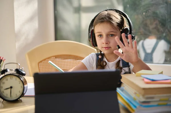 Distance learning. Online education. Langiage courses. Caucasian schoolgirl, a first grader wearing wireless audio headset sits at home at a desk with digital tablet studies at school online