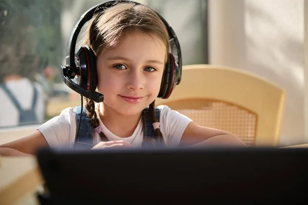Close-up portrait of a charming Caucasian little girl, first grader, schoolgirl in audio headset, sweetly smiles looking at camera while watching video lesson on laptop. Homeschooling. Back to school