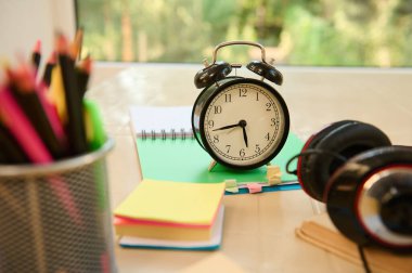 Still life. Focus on a black alarm clock on open notepad with school supplies, audio headset and stationery on blurred foreground. Back to school. Education. Teachers day concept