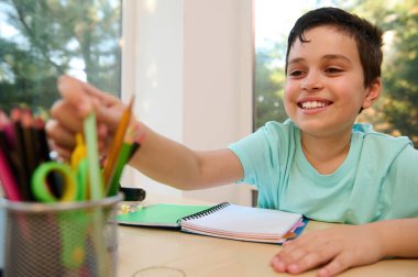 Portrait of an adorable school child, Caucasian boy smiling a cheerful toothy smile while taking out a pen from pencil holder. Homeschooling. Back to school. Knowledge and education concept. Lifestyle