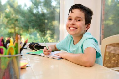 Adorable Caucasian child in bright blue t-shirt smiles looking at camera while making notes on copybook, siiting at a table with pencil holdre and school supplies on the foreground. Back to school