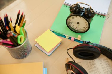 Still life. View from above with focus on a black alarm clock on open notepad with school supplies, audio headset and stationery on blurred foreground. Back to school. Education. Teachers day concept