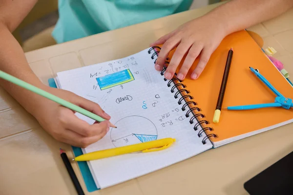 View from above of a math copybook, and a hand of a schoolboy holding a pencil solving problems in an online geometry lesson. New knowledge. Back to school. New semester of an academic year