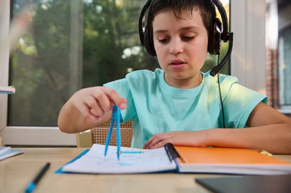 Portrait of an adorable schoolboy in audio headset, solving math problems using a compass to draw a circle in a notebook during an online geometry lesson. Back to school on new semester academic year
