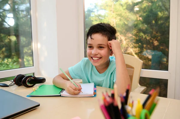 Happy Caucasian schoolboy, student of primary educational establishment, smiling a cheerful toothy smile while doing homework. Knowledge. Online Education. Back to school. New academic year semester.