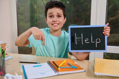 Adorable puzzled schoolboy holds a blackboard with the inscription HELP, points his finger at a notebook with examples of math and geometry, asks for help in solving problems. Difficulties in learning