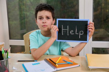 Puzzled schoolboy points with pencil on chalkboard with inscription Help, sitting at table with books and school supplies, asks for help on solving maths geometry problems. New academic year semester