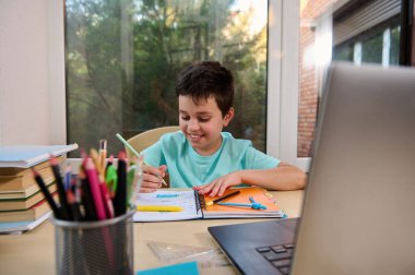 Front view of a cheerful happy schoolboy doing homework, learning geometry sitting at laptop during online lesson. Homeschooling, online education. Using internet for studying across the distance