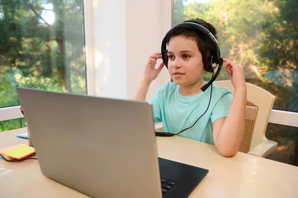 Handsome schoolboy wearing audio headset, sitting at a desk and using laptop watching broadcasted video lessons while studying remotely in an online school
