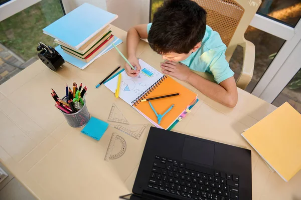 Top view. School boy solves maths and geometry examples, sitting at a table with school supplies, measuring rulers, stacked textbooks, black alarm clock and laptop, broadcasting an online video lesson