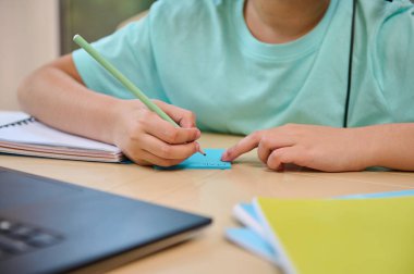 Cropped view of a schoolchild during a video conference with the teacher in virtual class, with selective focus on his hands holding a pencil and making notes during an online lesson on the laptop