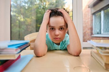 Portrait of Caucasian stunning smart boy, elementary school student holding his head and looking at camera. School supplies and textbooks are on the tabel. Online education and homeschooling concept