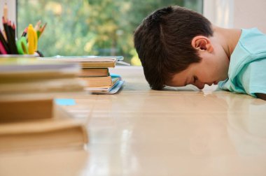 Tired bored schoolboy, primary student in online school, sitting at desk, leaning on the table with his forehead, having difficulty adapting to the new academic year semester. Homeschooling