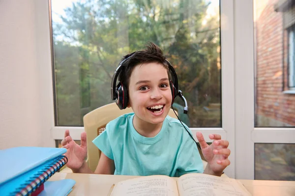 Cheerful schoolboy in headphones, smiles looking at camera while doing literature homework. Erudition. Back to school. Open to new knowledge in new semester of the new academic year. Online education