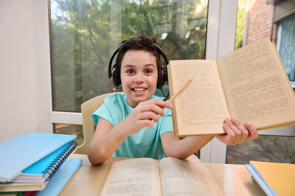 Front portrait of smart student of online elementary school in headphones, points with pencil at blurred text in textbook, looks at camera and confidently answers teachers questions during a lesson