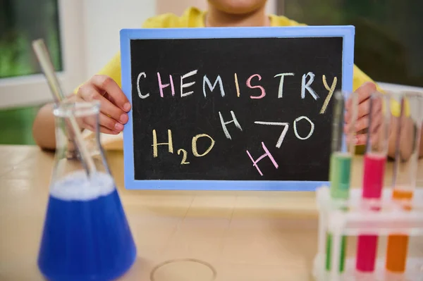 Blackboard with written inscription Chemistry, made with chalk in the hands of a schoolboy chemist, sitting at a desk with flask and test tubes filled with chemical liquids and reagents on the tripod