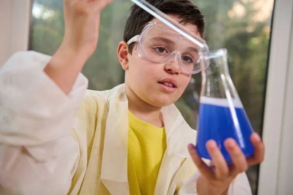 Smart schoolboy, scientist chemist in safety goggles and a white lab coat, dripping a reagent into a flat-bottomed flask with blue chemical liquid while doing a chemistry experiment in a school lab