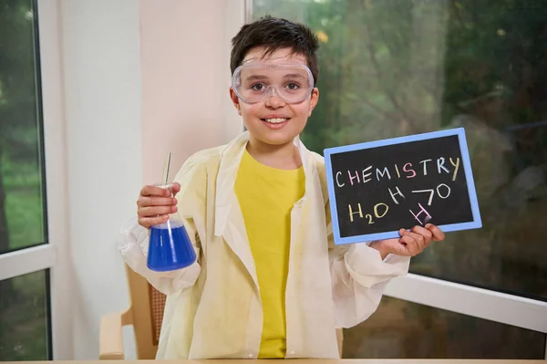 Little scientist chemist in safety goggles and white lab coat, holding a flask with blue chemical liquid and blackboard with the inscription Chemistry and the water formula written with colored chalks